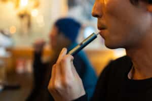 Close-up of a man smoking an E-cigarette in a bar with an out-of-focus person in the background also smoking an electronic cigarette.